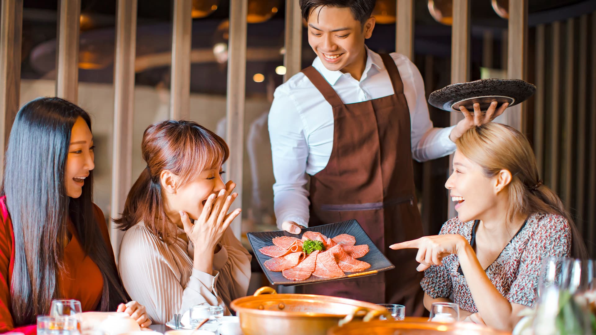 Nhân viên phục vụ nhà hàng Nhật tươi cười bưng đĩa thịt bò thái lát mời bàn khách đang trò chuyện vui vẻ quanh nồi lẩu đồng