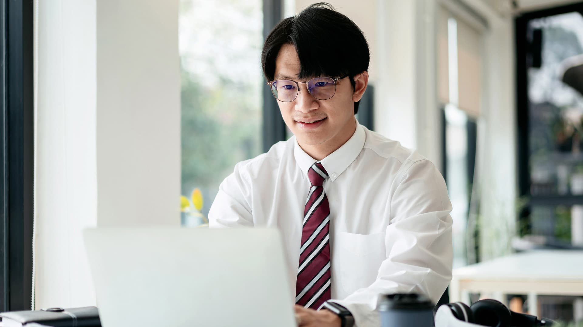Kỹ sư trẻ đeo kính, sơ mi cà vạt, làm việc bên laptop trong văn phòng công ty Nhật